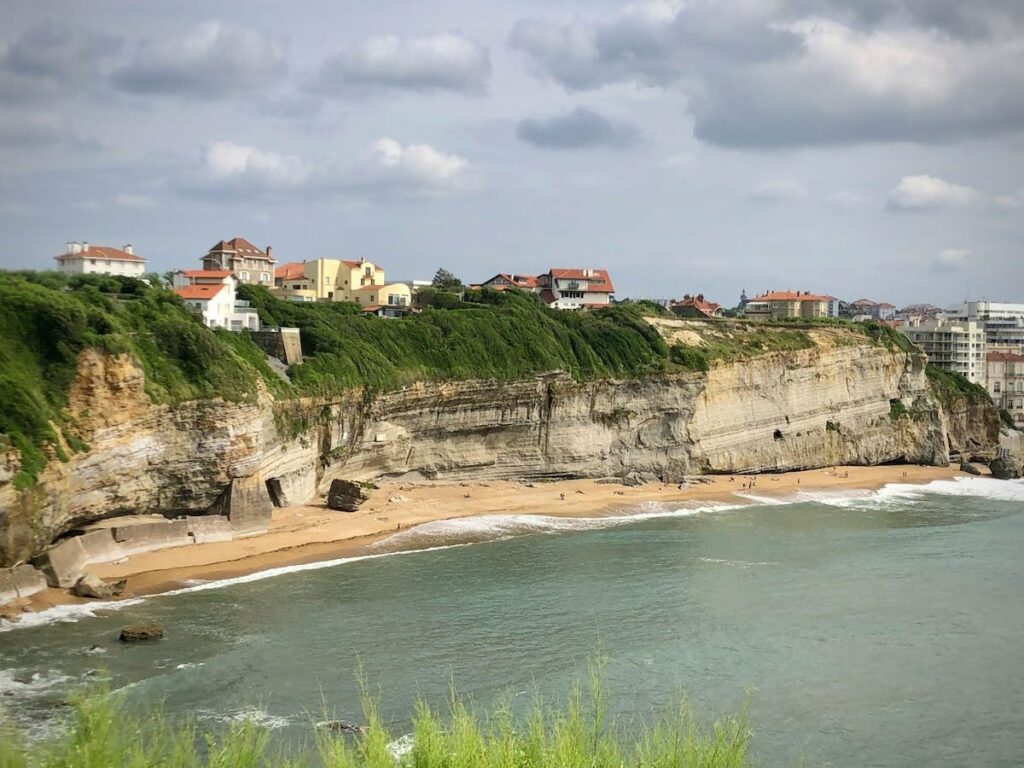 Cliffside houses overlooking a sandy beach and calm blue sea under a cloudy sky, with green vegetation atop the cliff edge.
