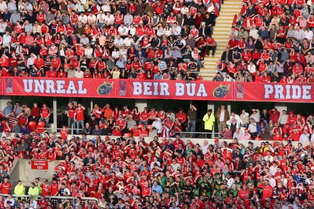 Crowd of red-clad sports fans filling stadium stands, with a long red banner across the upper level reading 'UNREAL BEIR BUA PRIDE'.