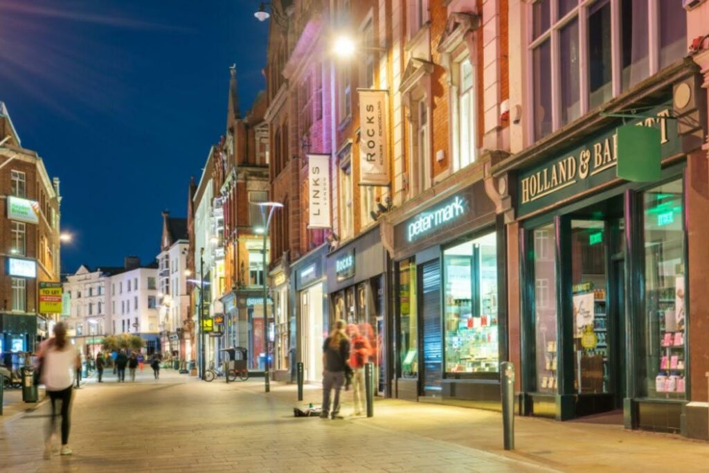 Night street scene with lit storefronts and pedestrians along a shopping street, showing signs like Peter Mark and Holland & Barrett.