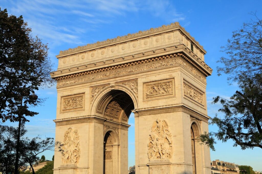 Front view of the Arc de Triomphe in Paris, with detailed reliefs and a bright blue sky above.