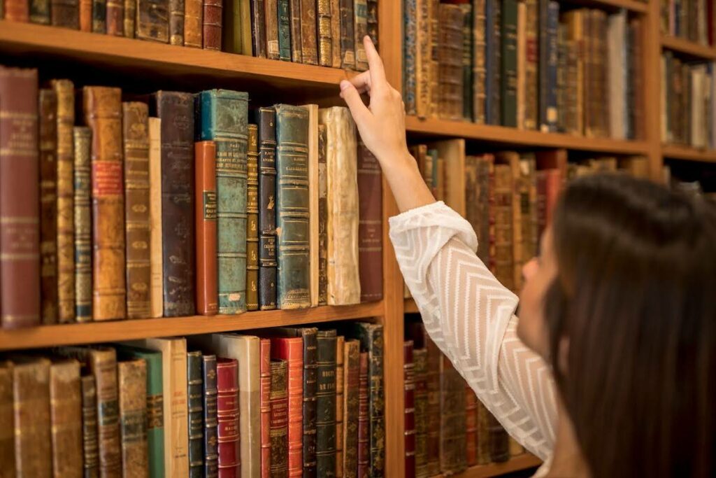 Person reaching for a book on a wooden library shelf filled with old volumes on the wall-length shelves in a library setting.