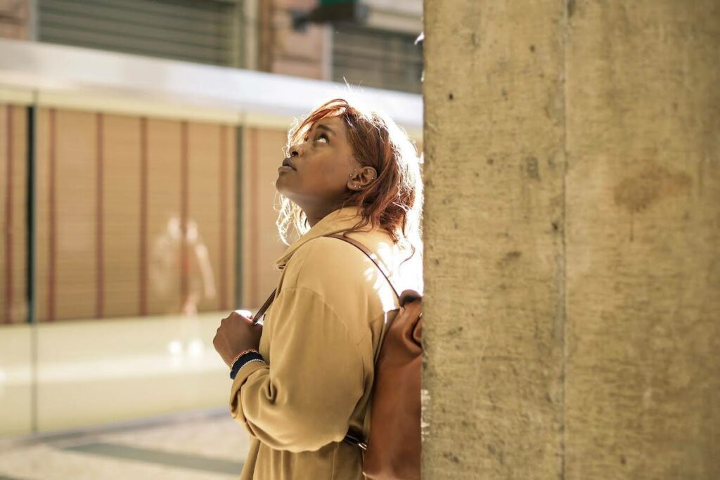 Woman in a tan coat with a brown backpack stands beside a concrete pillar, looking upward in an indoor urban space.