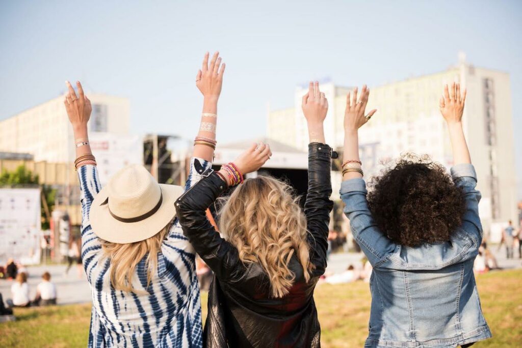 Three women with arms raised in celebration at an outdoor city park festival (back view).