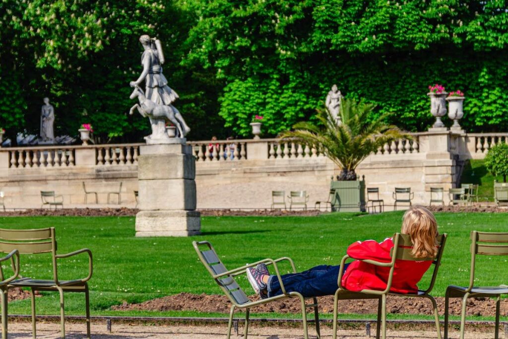 Person in a red jacket relaxing on a metal chair in a formal garden with statues and green hedges behind