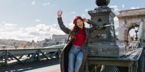 Woman in a red turtleneck and plaid coat sits on a bridge structure, smiling with a cup raised in one hand and a red beret on, sunny day.