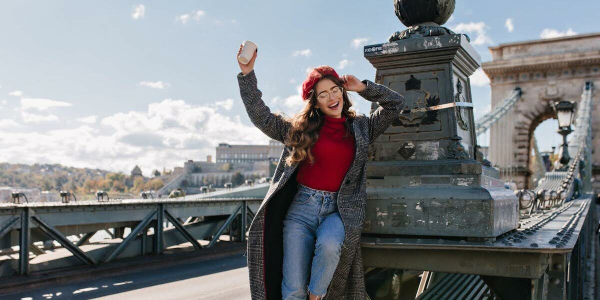 Woman in a red turtleneck and plaid coat sits on a bridge structure, smiling with a cup raised in one hand and a red beret on, sunny day.