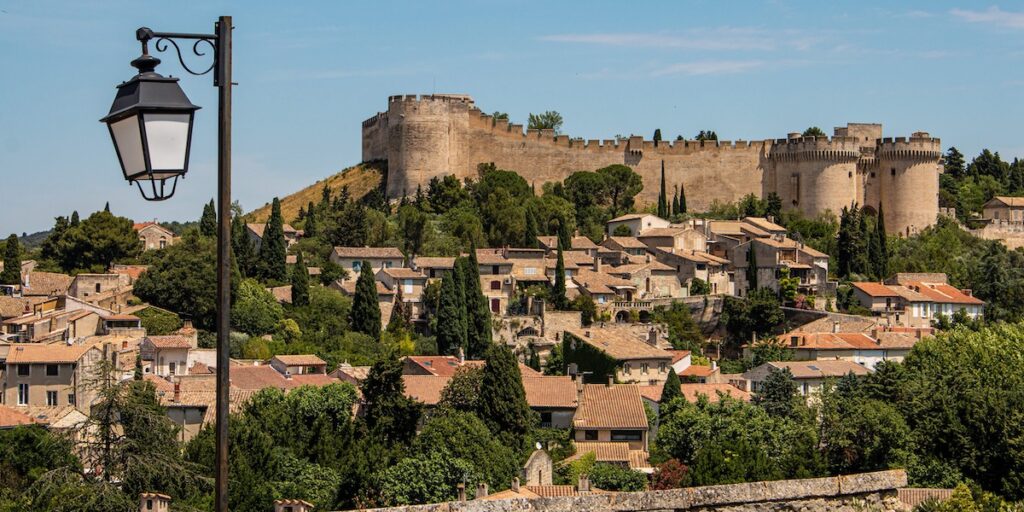 Medieval fortress wall and towers on a hill overlooking a terracotta-roofed village, with a vintage street lamp in the foreground.