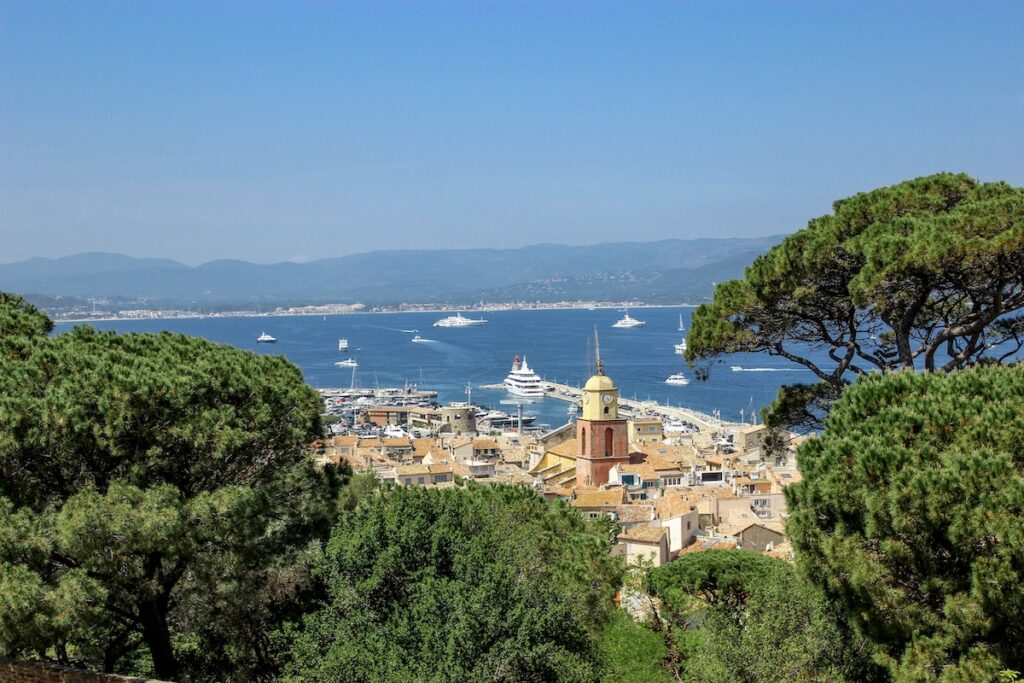 Harbor town with terracotta rooftops, a yellow clock tower, and boats in the blue bay framed by green trees.