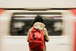 Person in a beige coat with a bright red backpack on a platform as a subway train whizzes by in motion blur.