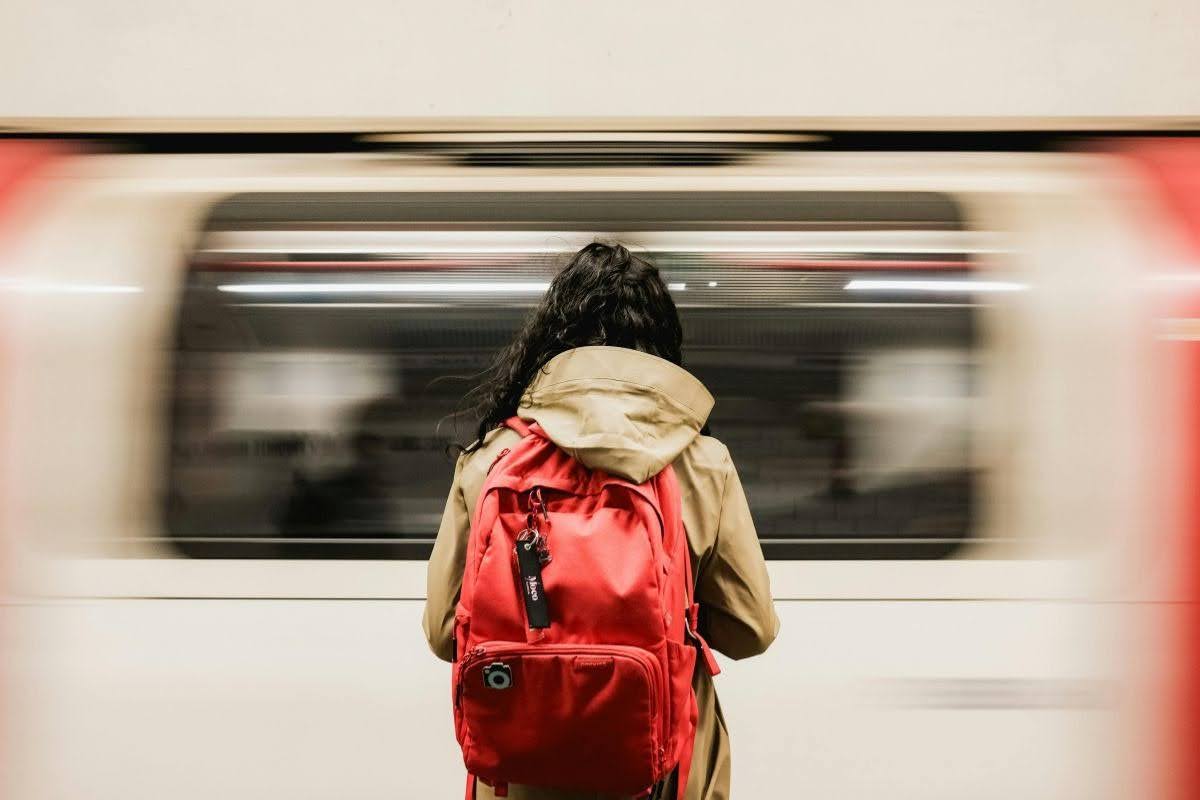 Person in a beige coat with a bright red backpack on a platform as a subway train whizzes by in motion blur.