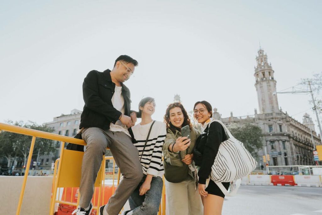 Four friends pose for a selfie on a sunny city street, beside yellow barriers and a tall historic building in the background.