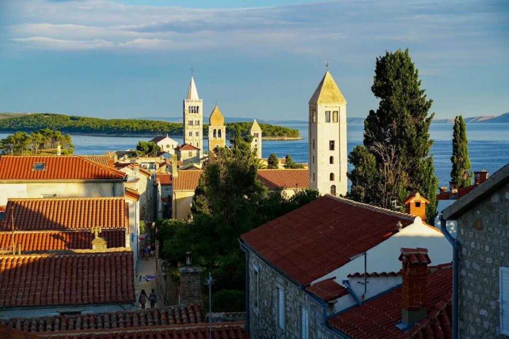 Coastal town with red-tiled roofs and stone towers overlooking a calm blue sea.