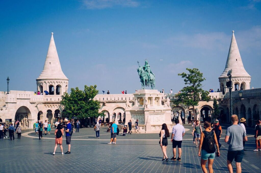 Tourists walk and stand in a sunny square at the Fisherman's Bastion in Budapest, with a bronze equestrian statue on a white pedestal and two cone-topped towers in the background.