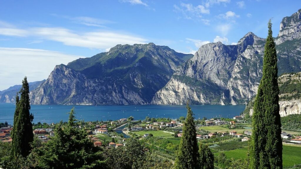 Blue lake nestled between dramatic mountains with a small village along the shore and tall evergreen trees in the foreground.