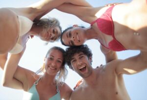 Five friends in swimwear form a circle above the camera, smiling and looking down on a sunny beach day.