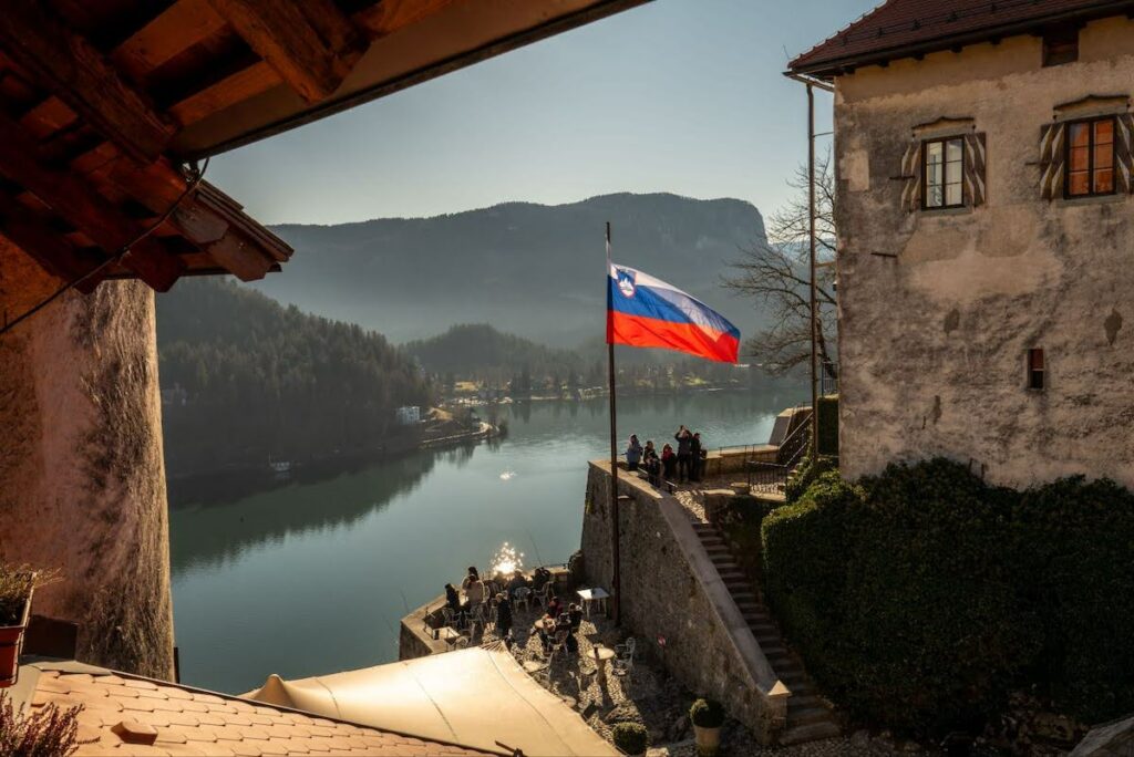 Slovenian flag over a stone terrace by a calm lake, with visitors gathered at tables and mountains in the background.