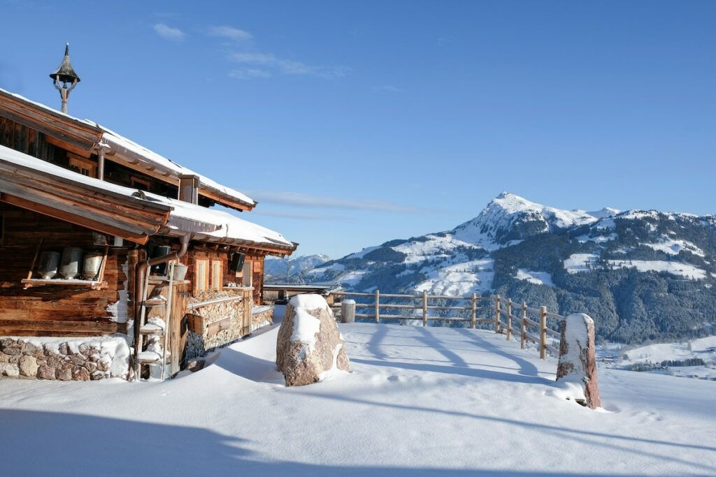 Snow-covered wooden chalet with stone foundation on a snowy slope, against distant alpine mountains under a blue sky.