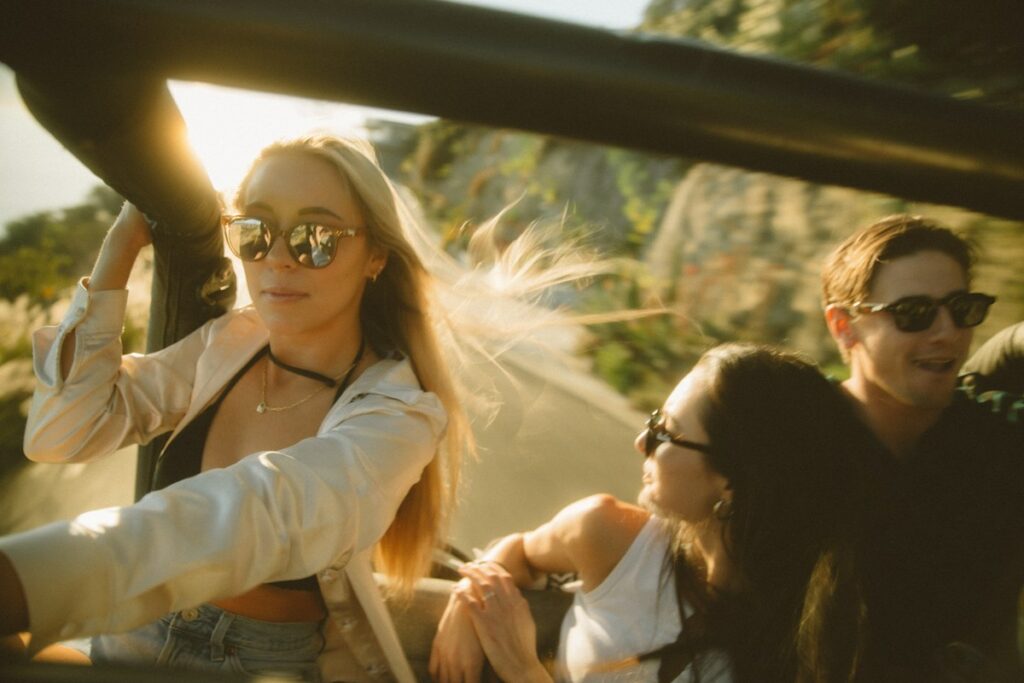Three friends in an open-top car on a sunny day, all wearing sunglasses and smiling.