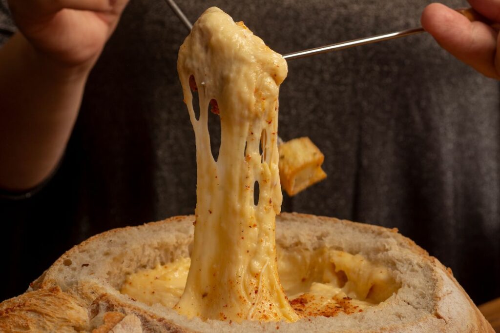 Melted cheese stringing from a fondue fork lifted over a bread bowl filled with cheese dip.