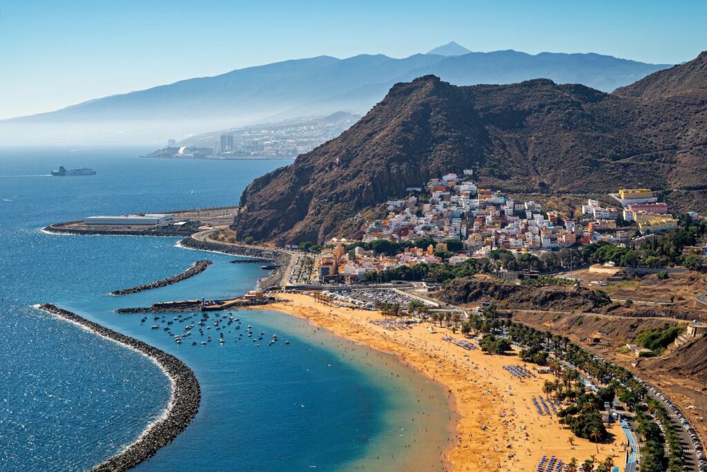 Aerial view of a coastal town built on a hillside with a sandy beach, blue sea, and distant mountains in the background.