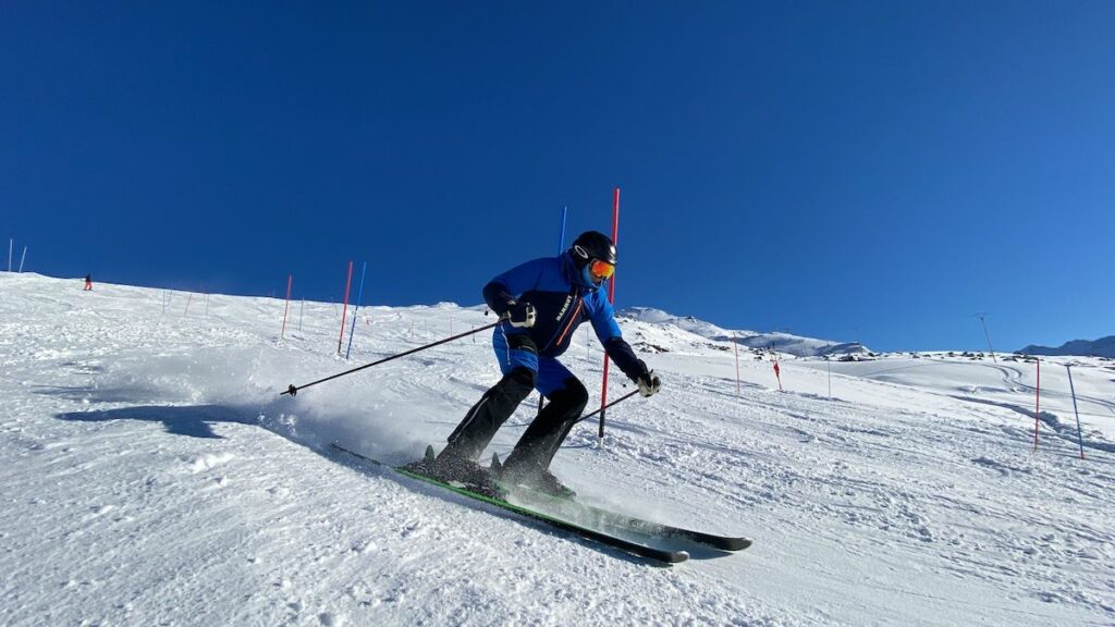 Skier in blue gear carving a turn on a snow-covered slope, navigating red and blue gates under a clear blue sky.