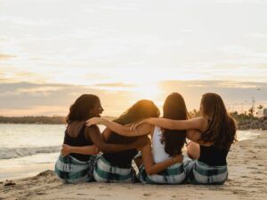 Four women sit on the sandy beach at sunset, arms around each other in a friendly circle.