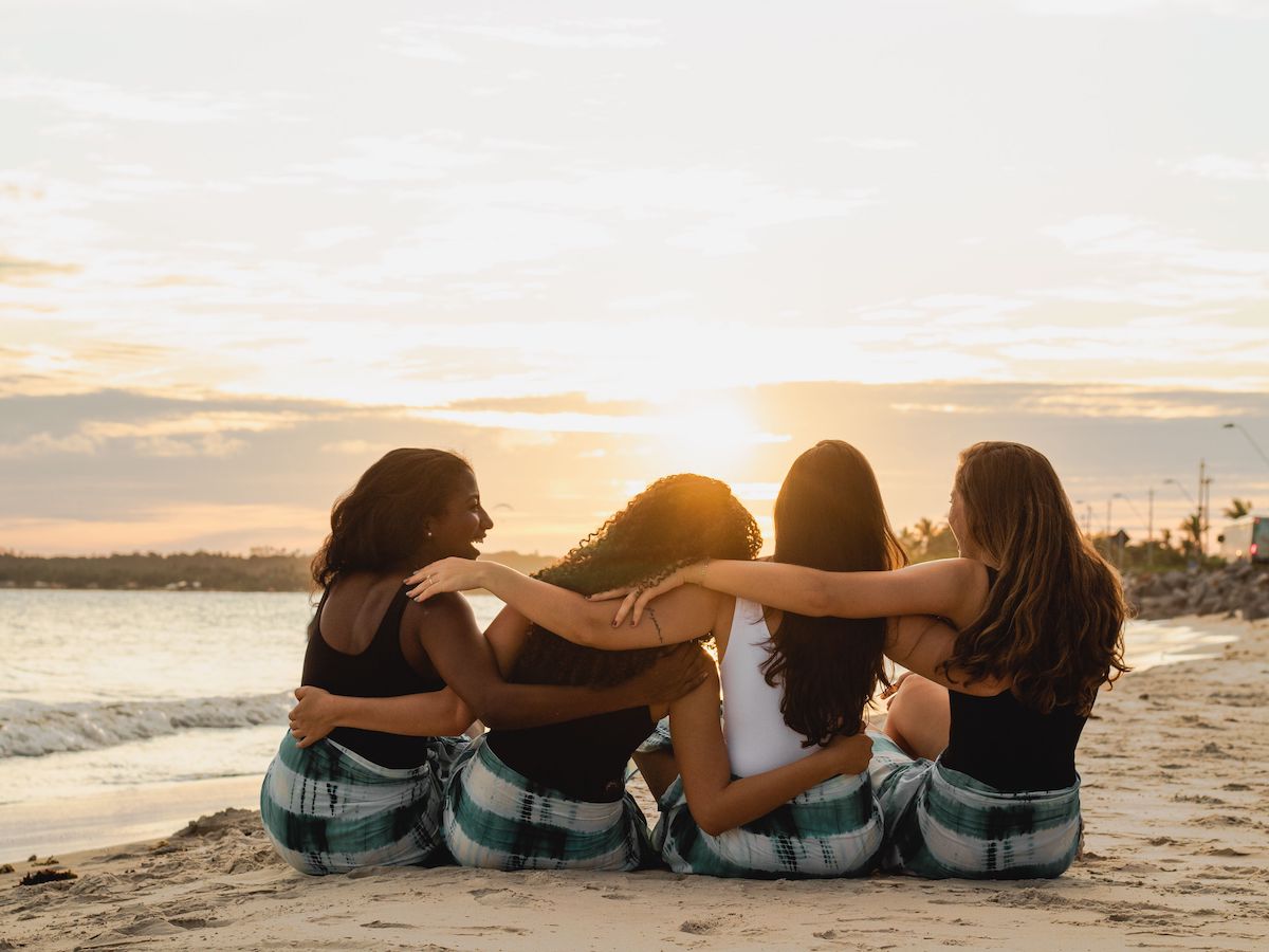 Four women sit on the sandy beach at sunset, arms around each other in a friendly circle.
