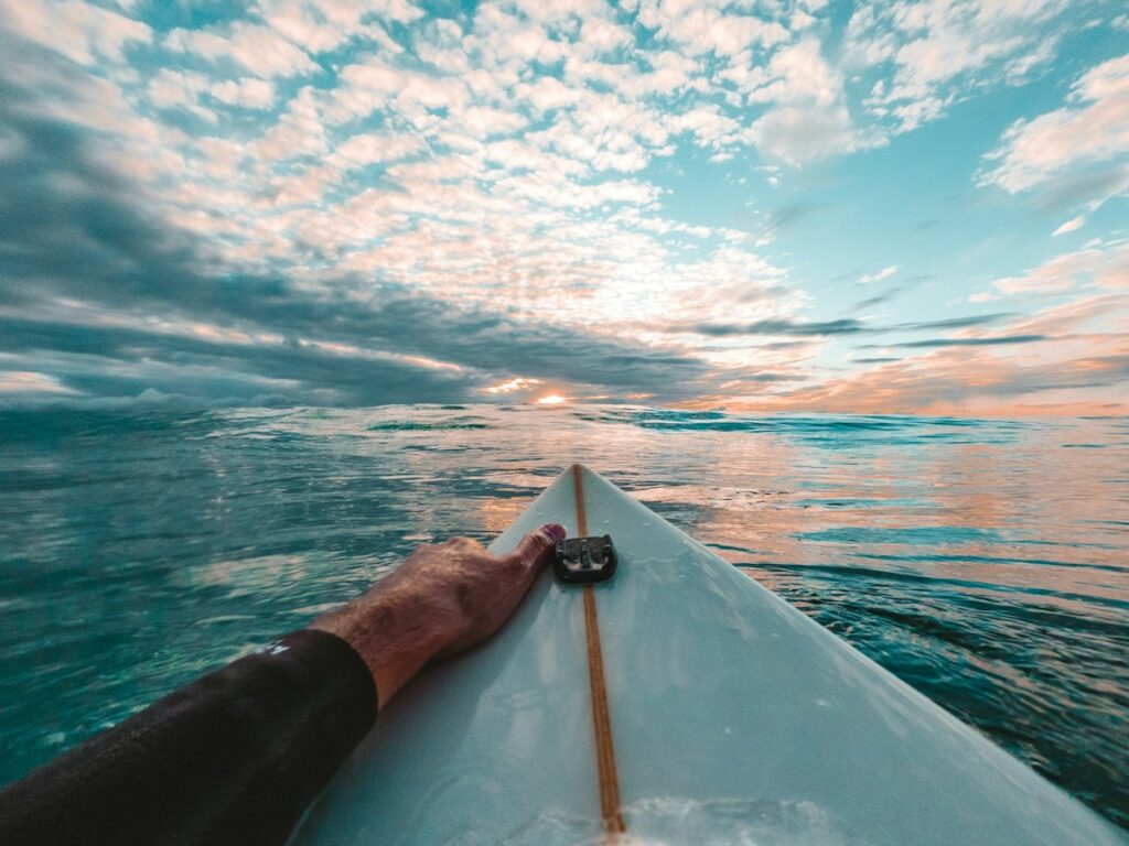 First-person view of a surfer's hand on a white surfboard as the sun sets over a calm ocean. Clouds glow orange and pink above turquoise water.