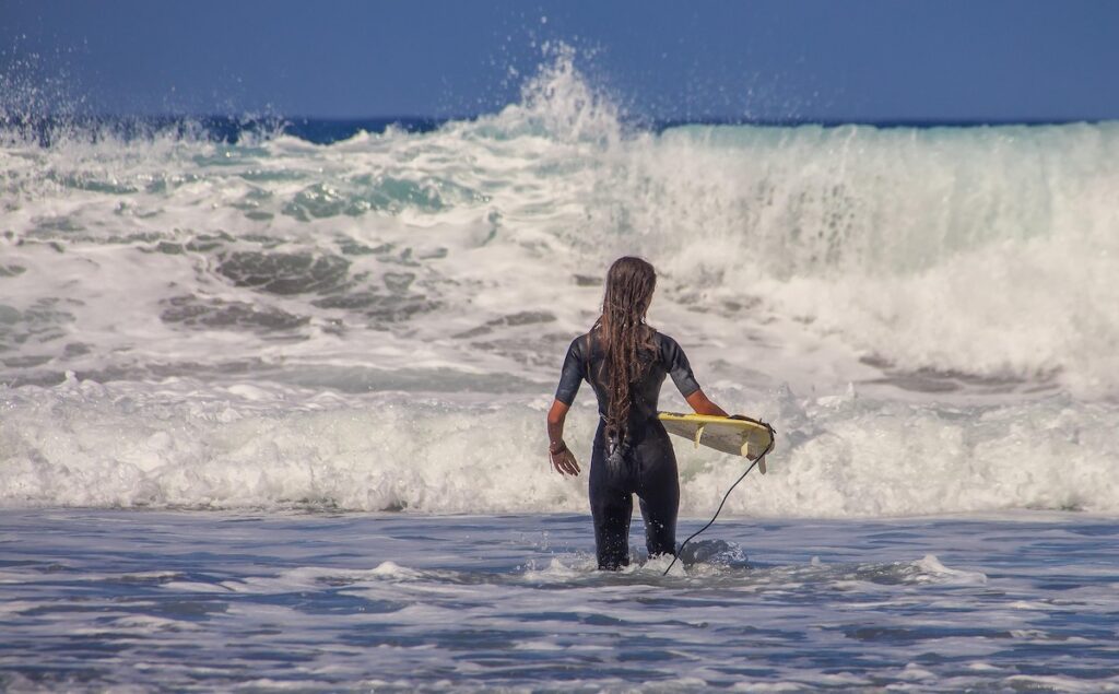 Surfer in a black wetsuit walks into the sea with a yellow surfboard under their arm, waves crashing nearby.