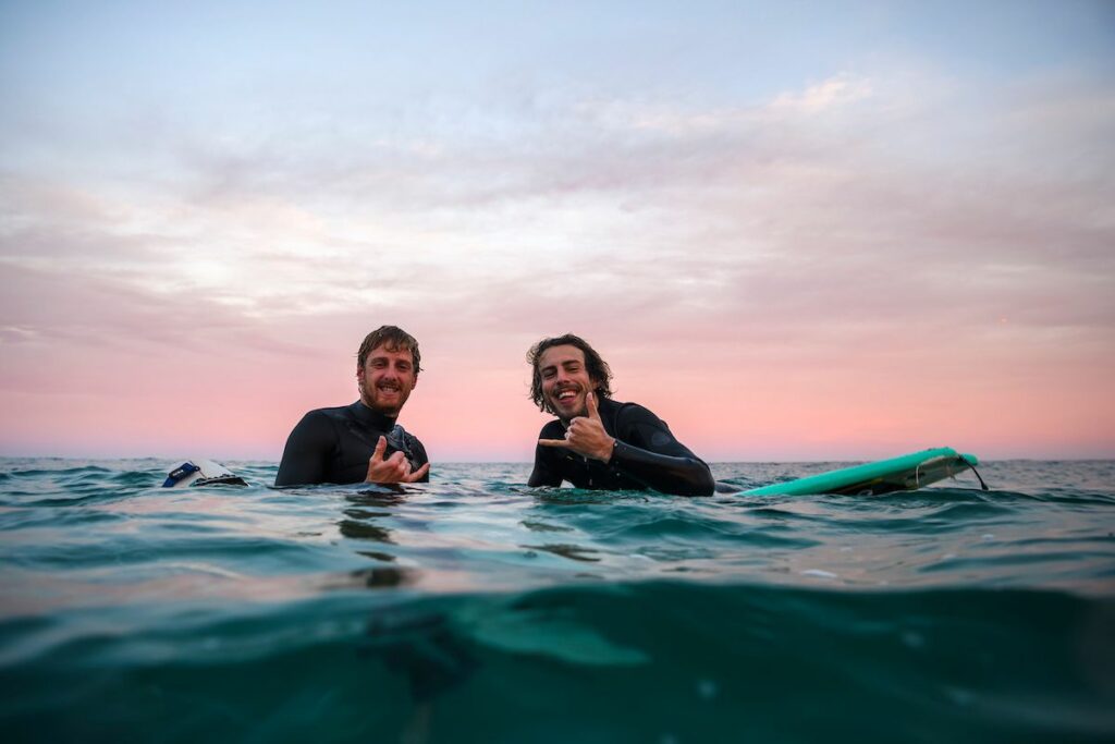 Two surfers in wetsuits smile and pose in the ocean at sunset, with a green surfboard nearby.