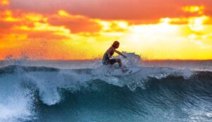 Surfer riding a breaking wave at sunset with spray around them and an orange sky behind.