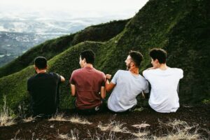 Four men sit with their backs to the camera on a rocky hillside, gazing over a green valley and distant hills.