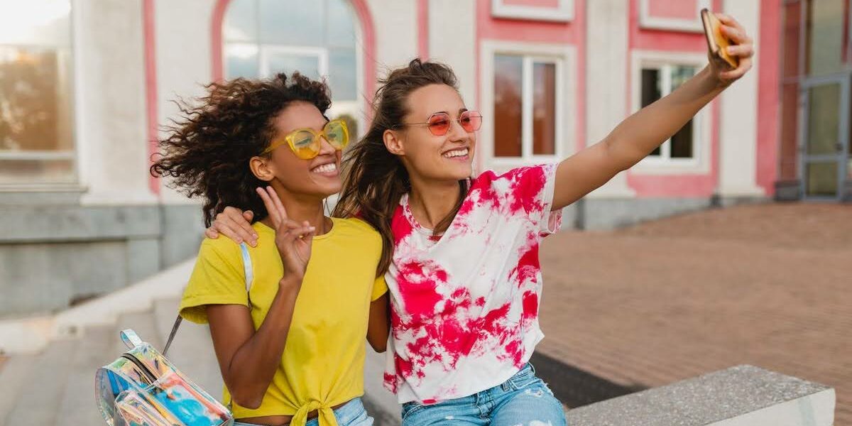 Two friends sit on a concrete ledge taking a cheerful selfie with a smartphone, wearing bright yellow and red tie-dye shirts and ripped denim shorts. One carries a clear bag with colorful items; sunny urban backdrop with pink buildings behind them.