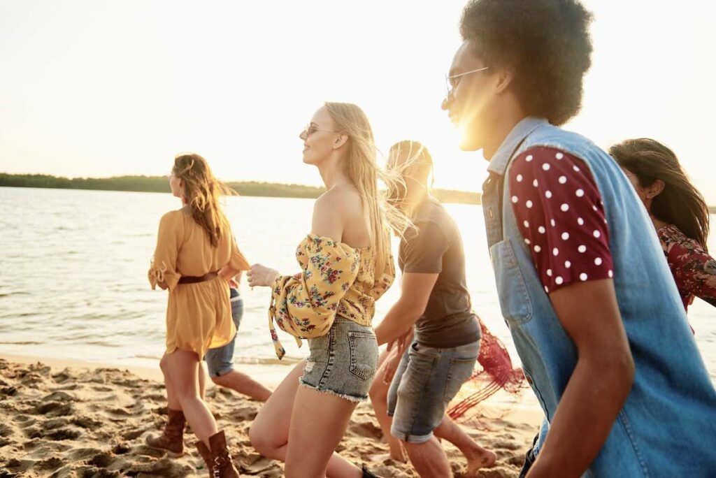 Group of friends walking along a sunlit beach at golden hour, chatting and laughing in casual summer outfits by the water.