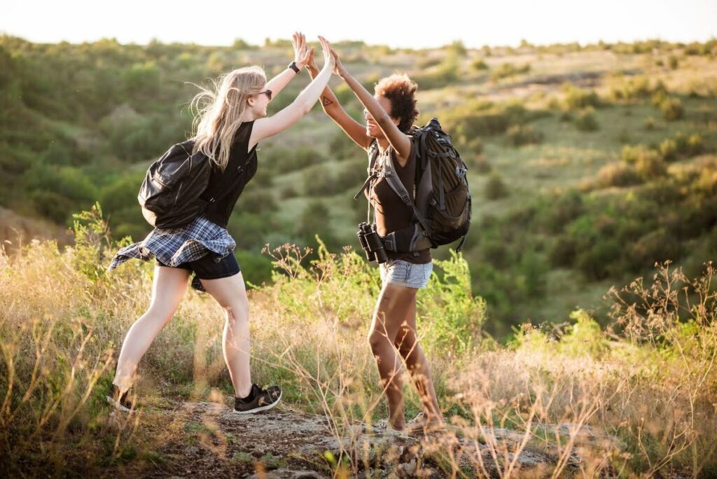 Two hikers on a sunlit hillside trail share a high-five, backpacks on and a sense of accomplishment.
