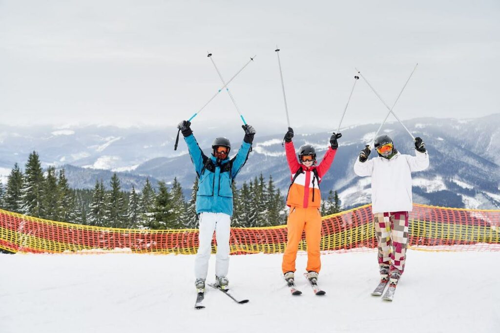 Three skiers in colorful outfits raise their poles in the air on a snowy slope with mountains in the background, wearing helmets and goggles.
