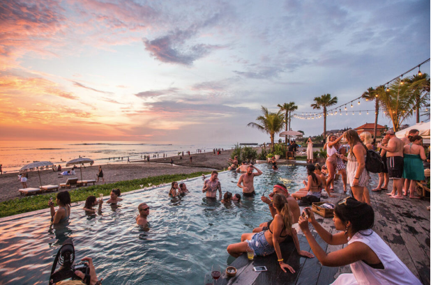 Sunset at a tropical beach resort with people in a pool and others lounging on a wooden deck under string lights and palm trees.