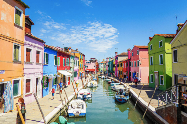 Colorful houses line a canal with small boats docked along both sides and a bright blue sky.